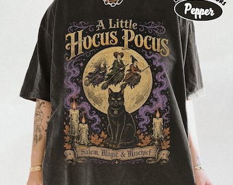 May include: A charcoal gray t-shirt featuring a graphic print with the text A Little Hocus Pocus and Salem, Magic & Mischief. The design includes three witches flying on brooms in front of a full moon, a black cat, and two lit candles.