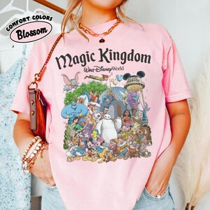 May include: Pink t-shirt with a graphic featuring various Disney characters and the text "Magic Kingdom Walt Disney World." The shirt also has the words "Comfort Colors Blossom" in a circular design.