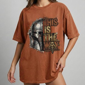 Może przedstawiać: Rdzawa koszulka z grafiką hełmu Mandalorianina i napisem "THIS IS THE WAY" w stylu vintage. Projekt zawiera metaliczne srebrne i złote akcenty, tworząc wygląd vintage.