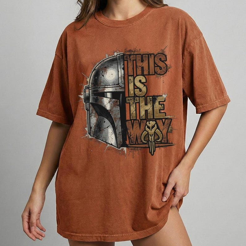 Może przedstawiać: Rdzawa koszulka z grafiką hełmu Mandalorianina i napisem "THIS IS THE WAY" w stylu vintage. Projekt zawiera metaliczne srebrne i złote akcenty, tworząc wygląd vintage.