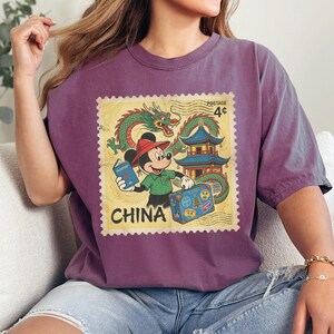 Op de afbeelding: Een paars T-shirt met een vintage postzegelontwerp met Mickey Mouse als toerist in China. De postzegel bevat een groene draak, een pagode en het woord "CHINA". Mickey houdt een paspoort en een koffer vast.