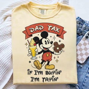 Puede incluir: Camiseta de color crema con un gr&aacute;fico de Mickey Mouse sosteniendo un batido y una rosquilla. El texto "DAD TAX" est&aacute; encima del personaje, con "If I'm buyin' I'm tryin'" debajo. La camiseta es de material suave.