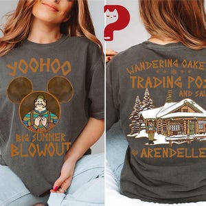 May include: Dark gray t-shirt featuring a cartoon image of a character with the text "Yoohoo Big Summer Blowout" on the front. The back of the shirt displays "Wandering Oaken's Trading Post and Sauna Arendelle" with a cabin graphic. The shirt is a casual wear item.