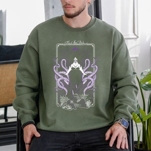 May include: A green sweatshirt with a graphic print of Ursula from The Little Mermaid. The graphic features Ursula in her octopus form with the text "The Sea Witch" above her.