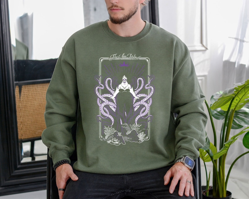 May include: A green sweatshirt with a graphic print of Ursula from The Little Mermaid. The graphic features Ursula in her octopus form with the text "The Sea Witch" above her.
