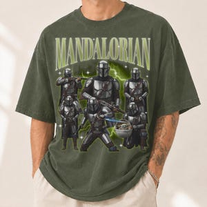 Puede incluir: Camiseta verde oliva con un gr&aacute;fico de varios Mandalorianos con armadura plateada. La palabra "MANDALORIAN" est&aacute; en verde claro, en un estilo vintage. El dise&ntilde;o incluye un beb&eacute; Yoda y rayos.