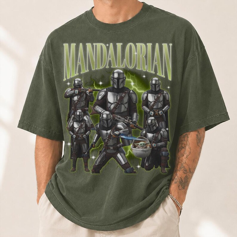 Puede incluir: Camiseta verde oliva con un gr&aacute;fico de varios Mandalorianos con armadura plateada. La palabra "MANDALORIAN" est&aacute; en verde claro, en un estilo vintage. El dise&ntilde;o incluye un beb&eacute; Yoda y rayos.