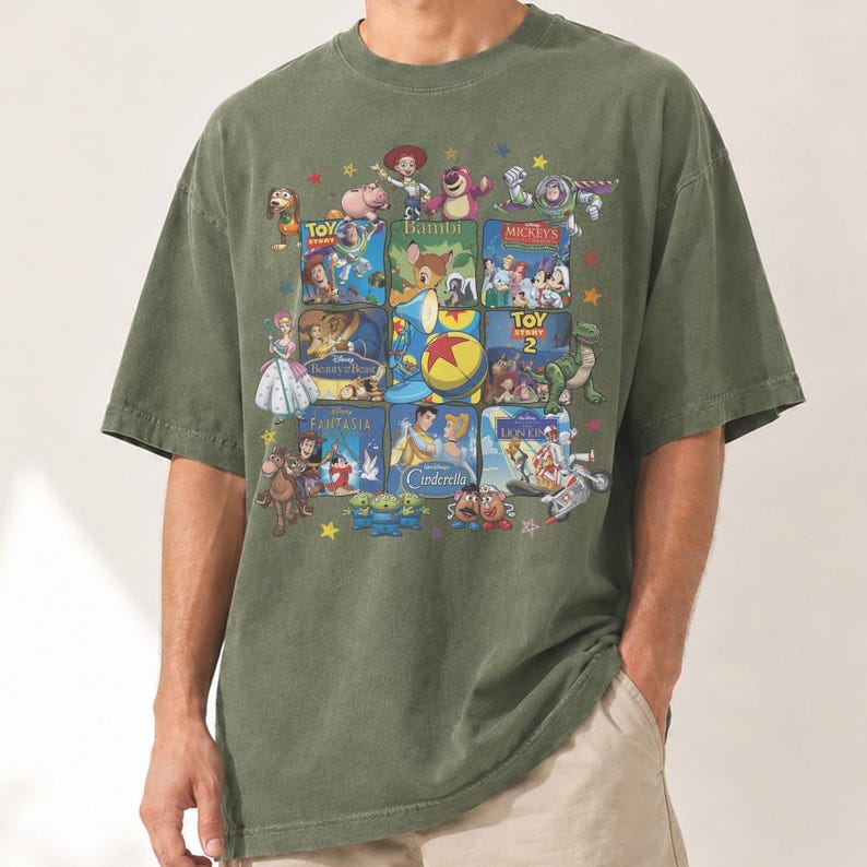 May include: Olive green t-shirt featuring a collage of classic Disney movie posters. The design includes images from "Toy Story," "Bambi," "Cinderella," and more, with a scattering of stars. The shirt has a relaxed fit.