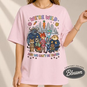 K&ouml;nnte beinhalten: Hellrosa T-Shirt mit einer Grafik mit Zeichentrickfiguren und dem Text "WE'RE WILD AND WE CAN'T BE TAMED". Das Design umfasst eine Stadtlandschaft, einen Donut, eine Karotte und Pfotenabdr&uuml;cke. Das Shirt ist von Comfort Colors.