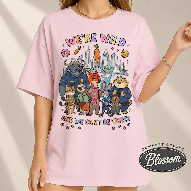 K&ouml;nnte beinhalten: Hellrosa T-Shirt mit einer Grafik mit Zeichentrickfiguren und dem Text "WE'RE WILD AND WE CAN'T BE TAMED". Das Design umfasst eine Stadtlandschaft, einen Donut, eine Karotte und Pfotenabdr&uuml;cke. Das Shirt ist von Comfort Colors.