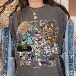 May include: Dark gray sweatshirt featuring a Halloween-themed graphic with Disney characters. The design includes Mickey and Minnie Mouse, Sully, Buzz Lightyear, and other characters in costumes, with the words "Hollywood Studios" and "Disney" visible.