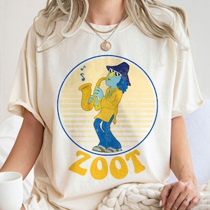 May include: A white t-shirt with a retro-style graphic of a blue character playing a yellow saxophone. The graphic is surrounded by a yellow circle with the word "ZOOT" in yellow letters.