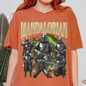Puede incluir: Camiseta de color naranja quemado con un gr&aacute;fico de los personajes de The Mandalorian. El dise&ntilde;o incluye varias figuras con armadura plateada, un efecto de rayo verde y la palabra "MANDALORIAN" en letras verdes.