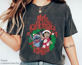 May include: Dark gray t-shirt featuring the phrase "Mele Kalikimaka" in red, with a wreath design. The wreath includes cartoon characters Stitch and Lilo, wearing Santa hats, surrounded by red flowers and green leaves.