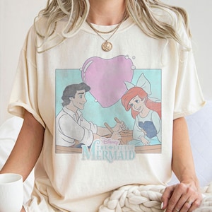 May include: A cream-colored t-shirt with a vintage-style graphic of Ariel and Prince Eric from Disney's The Little Mermaid. The graphic features a heart-shaped bubble and the text "Disney The Little Mermaid".