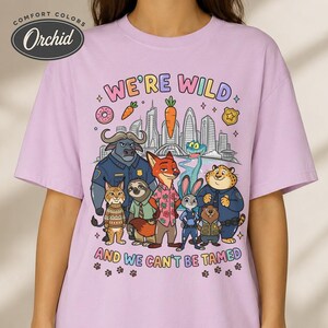 K&ouml;nnte beinhalten: Helllila T-Shirt mit farbenfroher Grafik mit Zeichentieren und dem Text "WE'RE WILD AND WE CAN'T BE TAMED". Das Shirt hat einen Rundhalsausschnitt und kurze &Auml;rmel.