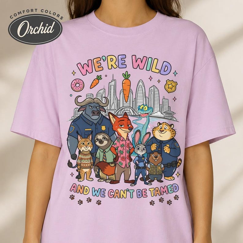 K&ouml;nnte beinhalten: Helllila T-Shirt mit farbenfroher Grafik mit Zeichentieren und dem Text "WE'RE WILD AND WE CAN'T BE TAMED". Das Shirt hat einen Rundhalsausschnitt und kurze &Auml;rmel.