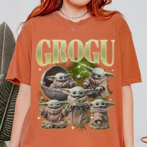 May include: Orange-brown t-shirt featuring a graphic of Grogu, also known as Baby Yoda, from The Mandalorian. The design includes multiple images of Grogu, a space pod, and the word "GROGU" in a green, glowing font.