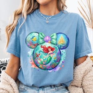 May include: A pale blue t-shirt with a colourful graphic of Ariel from The Little Mermaid. The design is in the shape of Mickey Mouse ears, with a bow and seashell accents. The graphic includes underwater scenes with fish, a crab, and a castle.