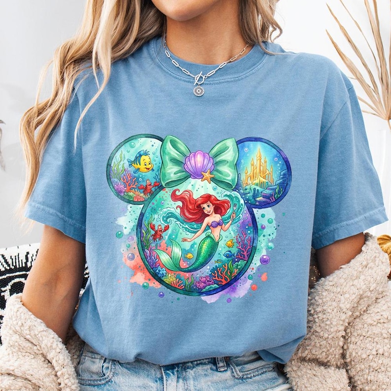 May include: A pale blue t-shirt with a colourful graphic of Ariel from The Little Mermaid. The design is in the shape of Mickey Mouse ears, with a bow and seashell accents. The graphic includes underwater scenes with fish, a crab, and a castle.