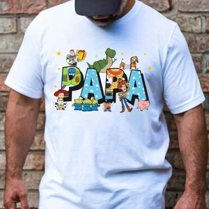 Puede incluir: Camiseta blanca con un gr&aacute;fico colorido de los personajes de Toy Story que deletrean "PAPA".