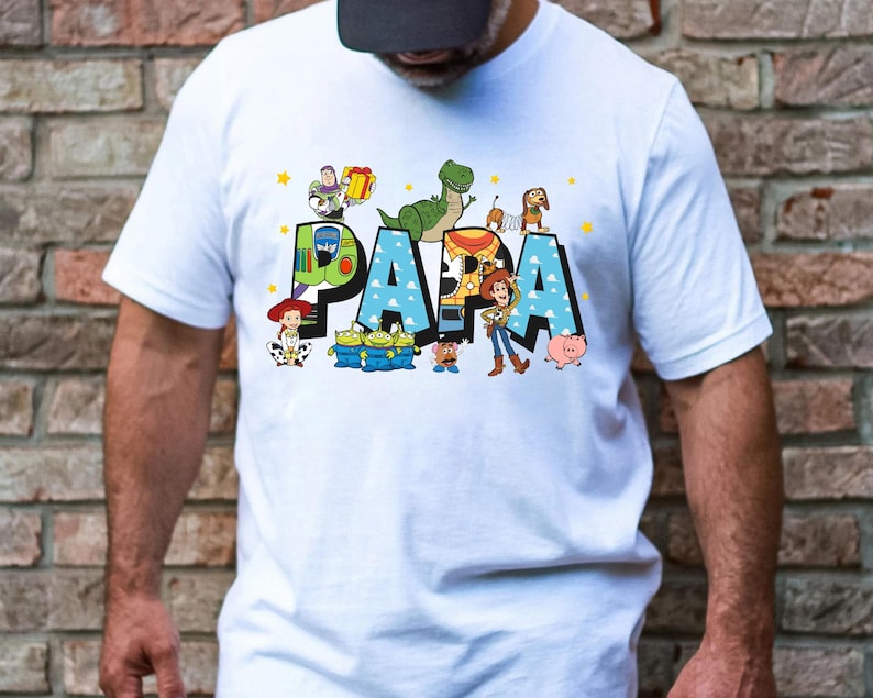 Puede incluir: Camiseta blanca con un gr&aacute;fico colorido de los personajes de Toy Story que deletrean "PAPA".