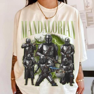 Puede incluir: Camiseta color crema con la palabra "MANDALORIAN" en verde, con un gr&aacute;fico de varias figuras mandalorianas con armadura plateada. Una figura sostiene a un beb&eacute; en una c&aacute;psula flotante. El dise&ntilde;o incluye rayos.