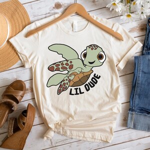 May include: A beige t-shirt with a cartoon sea turtle graphic and the text "Lil Dude".