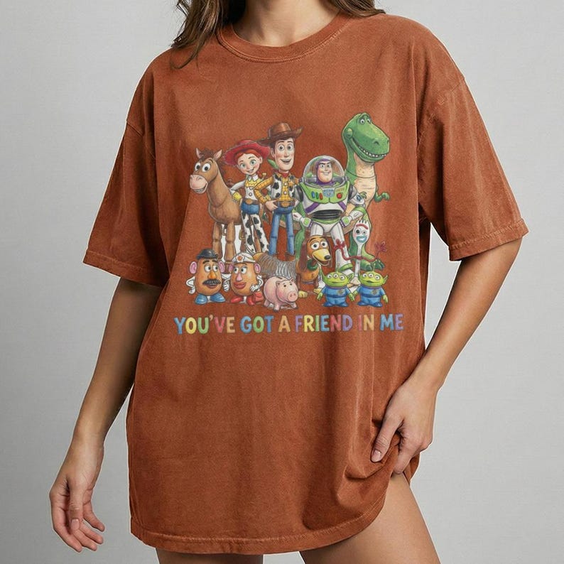 May include: A brown t-shirt featuring a colorful graphic of characters from the animated film "Toy Story." The graphic includes Woody, Buzz Lightyear, and other characters, with the text "YOU'VE GOT A FRIEND IN ME" in a rainbow font.