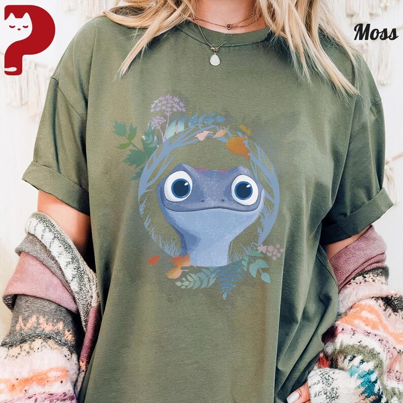 Puede incluir: Camiseta verde musgo con una ilustraci&oacute;n de dibujos animados de una salamandra azul con ojos grandes, rodeada por una corona floral. El dise&ntilde;o est&aacute; sobre un fondo de hojas y flores. La camiseta tiene mangas enrolladas.