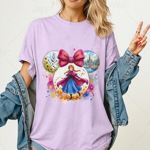 Puede incluir: Camiseta lila claro con un gr&aacute;fico colorido de un personaje de una popular pel&iacute;cula de animaci&oacute;n. El dise&ntilde;o incluye un gran lazo rosa e im&aacute;genes circulares de un castillo, un mu&ntilde;eco de nieve y el personaje con un vestido azul.