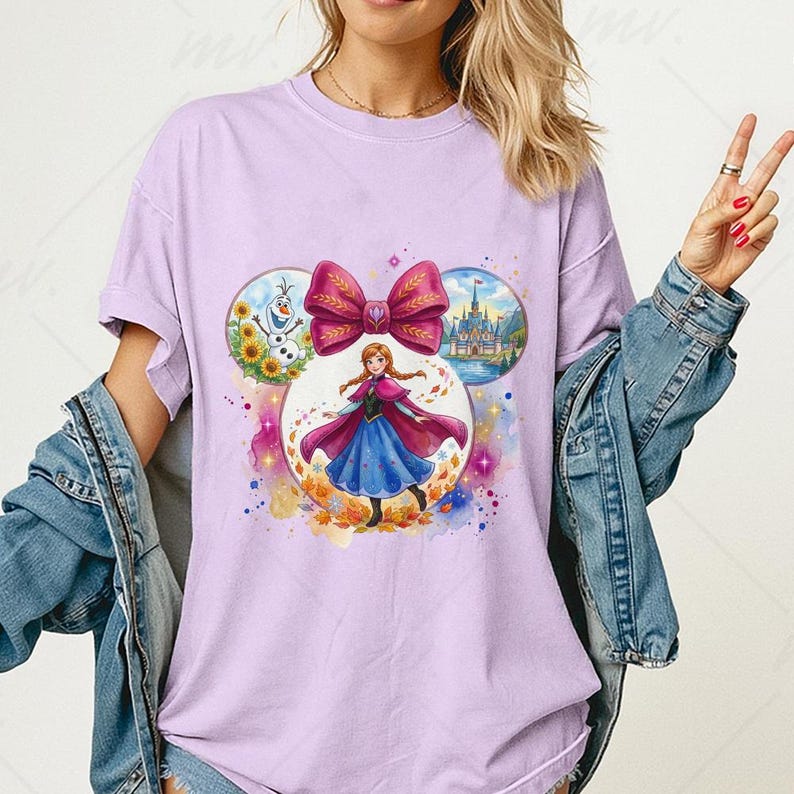 Puede incluir: Camiseta lila claro con un gr&aacute;fico colorido de un personaje de una popular pel&iacute;cula de animaci&oacute;n. El dise&ntilde;o incluye un gran lazo rosa e im&aacute;genes circulares de un castillo, un mu&ntilde;eco de nieve y el personaje con un vestido azul.