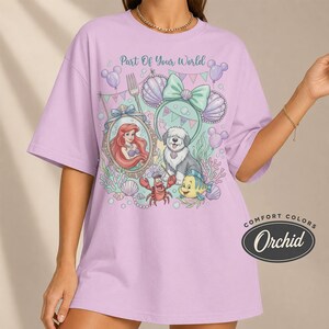 Może przedstawiać: Jasnofioletowa koszulka z grafiką przedstawiającą Ariel, psa i inne postacie z Małej Syrenki. Tekst "Part Of Your World" znajduje się nad grafiką. Koszulka ma etykietę Comfort Colors Orchid.