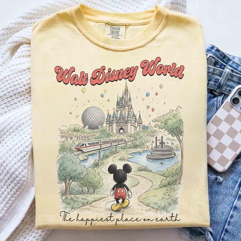 May include: Yellow Comfort Colors t-shirt featuring a colorful illustration of Walt Disney World. The design includes the iconic Cinderella Castle, Spaceship Earth, a monorail, and Mickey Mouse. Text reads "Walt Disney World" and "The happiest place on earth."