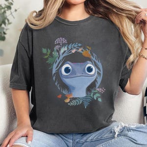 Puede incluir: Camiseta gris oscuro con un dise&ntilde;o de personaje de lagarto azul, rodeado por una corona de hojas y flores. La ilustraci&oacute;n est&aacute; centrada, ofreciendo una apariencia caprichosa y con tem&aacute;tica de la naturaleza.