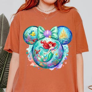 May include: Rust-coloured t-shirt featuring a colourful graphic of Ariel from The Little Mermaid. The design incorporates Mickey Mouse ears with underwater scenes, a bow, and a seashell. The artwork includes vibrant blues, greens, and purples.