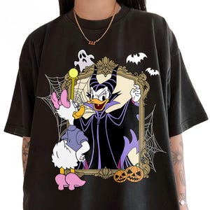 May include: Black t-shirt featuring a Halloween-themed graphic. The design includes Daisy Duck dressed as a witch, and a framed image of Maleficent. Bats, ghosts, spiderwebs, and pumpkins complete the spooky scene.