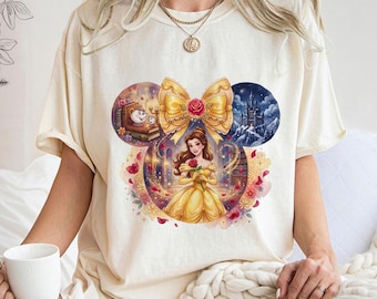 Puede incluir: Camiseta color crema con un gráfico colorido de Bella de La Bella y la Bestia. El diseño incluye un gran lazo amarillo, una rosa e ilustraciones del castillo, libros y personajes. La camiseta tiene mangas cortas y un corte holgado.