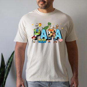 Puede incluir: Una camiseta beige con un gr&aacute;fico azul, verde y amarillo de personajes de Toy Story que deletrean "PAPA".