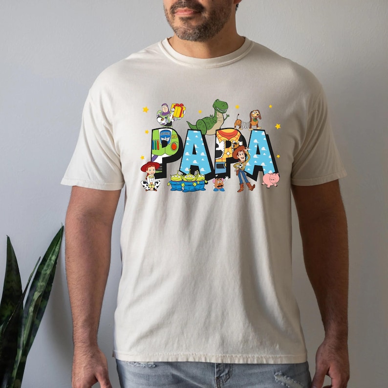 Puede incluir: Una camiseta beige con un gr&aacute;fico azul, verde y amarillo de personajes de Toy Story que deletrean "PAPA".