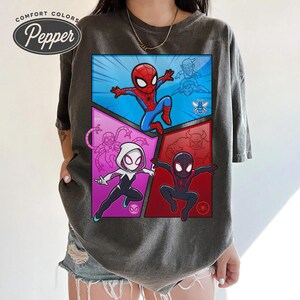 May include: Dark gray t-shirt featuring a comic book style graphic of Spider-Man, Spider-Gwen, and Miles Morales. The graphic is divided into four panels with a blue, red, and purple color scheme. The shirt has the text "Comfort Colors Pepper" at the top.