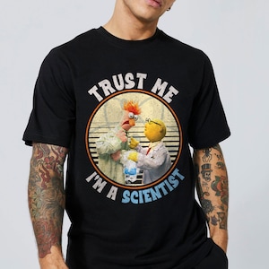 May include: Black t-shirt featuring a graphic of two Muppet characters in lab coats with the text "TRUST ME I'M A SCIENTIST" in a circular design. The design has a vintage, distressed look.