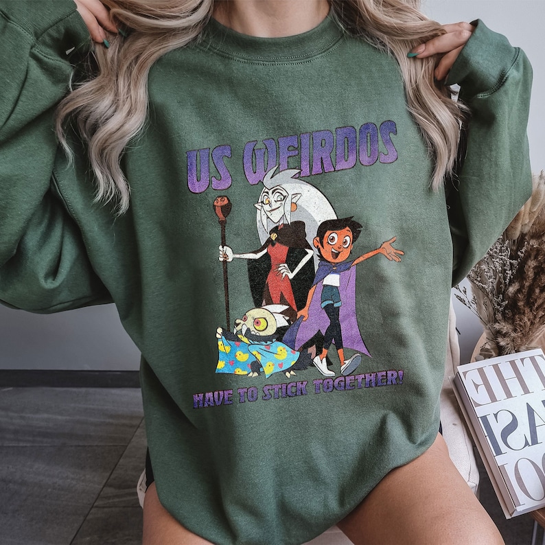 Op de afbeelding: Groen sweatshirt met een grafische print van personages uit de animatieserie "The Owl House". De tekst "US Weirdos Have to Stick Together!" staat op het sweatshirt gedrukt.