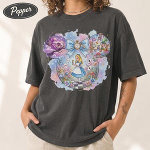 May include: Dark gray t-shirt featuring a colorful illustration of Alice in Wonderland within a Mickey Mouse ear outline. The design includes Alice, the Cheshire Cat, and other characters from the story. The shirt has a relaxed fit.