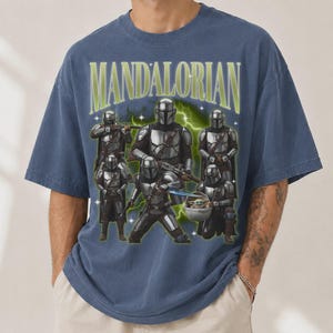 Puede incluir: Camiseta azul con un dise&ntilde;o de personajes Mandalorianos y el texto "MANDALORIAN" en verde y blanco. El dise&ntilde;o incluye m&uacute;ltiples figuras con armadura plateada, con un efecto de rayo verde.