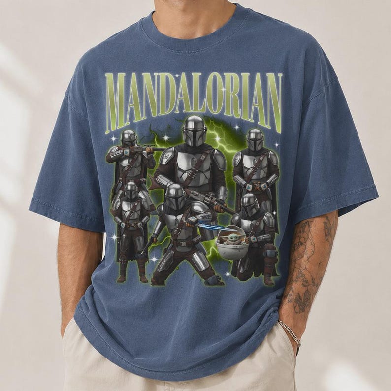 Puede incluir: Camiseta azul con un dise&ntilde;o de personajes Mandalorianos y el texto "MANDALORIAN" en verde y blanco. El dise&ntilde;o incluye m&uacute;ltiples figuras con armadura plateada, con un efecto de rayo verde.