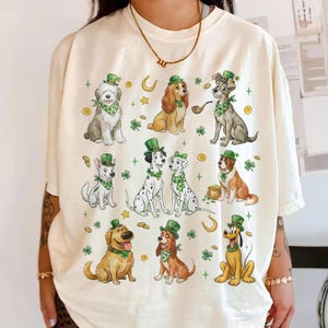 May include: Cream-colored t-shirt featuring cartoon dogs in St. Patrick's Day attire. The dogs are wearing green hats and bandanas, surrounded by shamrocks, horseshoes, and gold coins. The design includes various dog breeds in a whimsical illustration.