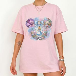 May include: A light pink t-shirt featuring a colorful graphic of Alice in Wonderland. The design is in the shape of Mickey Mouse ears, with Alice in the center, surrounded by iconic characters and imagery from the story.