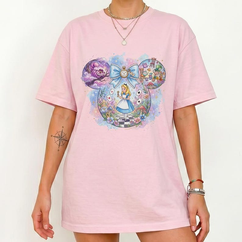 May include: A light pink t-shirt featuring a colorful graphic of Alice in Wonderland. The design is in the shape of Mickey Mouse ears, with Alice in the center, surrounded by iconic characters and imagery from the story.
