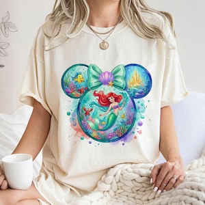 May include: Cream-coloured t-shirt with a colourful graphic of Ariel from The Little Mermaid. The design is in the shape of Mickey Mouse ears, with Ariel in the centre, surrounded by sea creatures and a castle. A large teal bow and seashell complete the design.